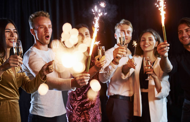 Having fun with sparklers. Group of cheerful friends celebrating new year indoors with drinks in hands. - Foto, Imagem