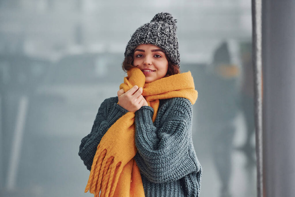 Beautiful cheerful girl in yellow scarf and in warm clothes standing indoors against background with reflections. - Foto, immagini