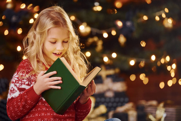 Cute little girl in red festive sweater reading book indoors at christmas holiday time. - Foto, Bild