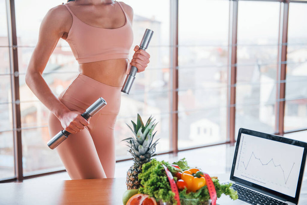 Female athlete in sportive wear holds dumbbells and standing near table with laptop and healthy food. - Foto, afbeelding
