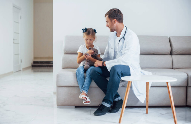 Male doctor in white uniform sits in the clinic with little girl. - Foto, Imagem