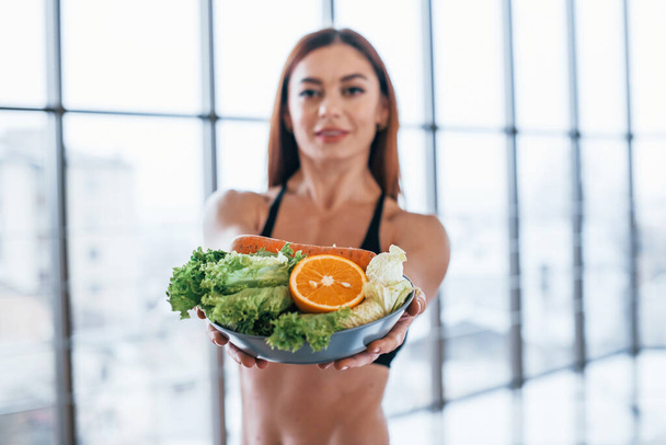 Cheerful sportive woman stands indoors with healthy food in hands. - Foto, imagen