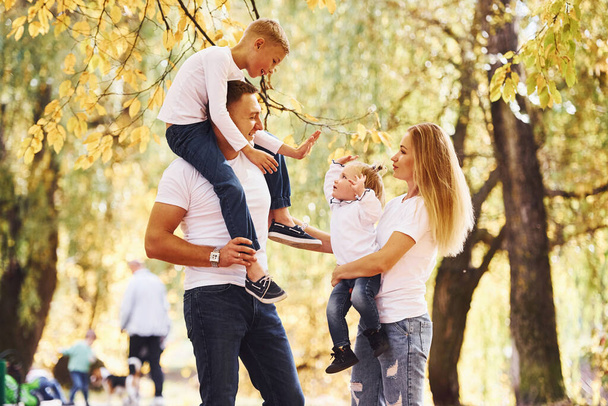 Mother and dad holds kids at shoulders and in hands. Cheerful young family have a walk in an autumn park together. - 写真・画像