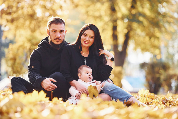 Cheerful family sits on the ground and having fun together with their child in beautiful autumn park. - Photo, Image