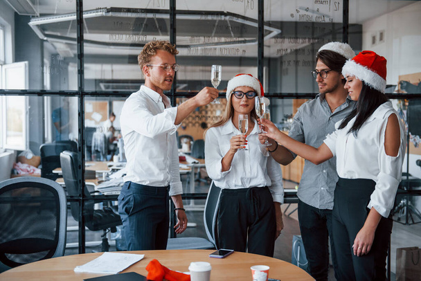 Employees in formal clothes, glasses with champagne and in christmas hats celebrating new year in the office. - Foto, Imagen