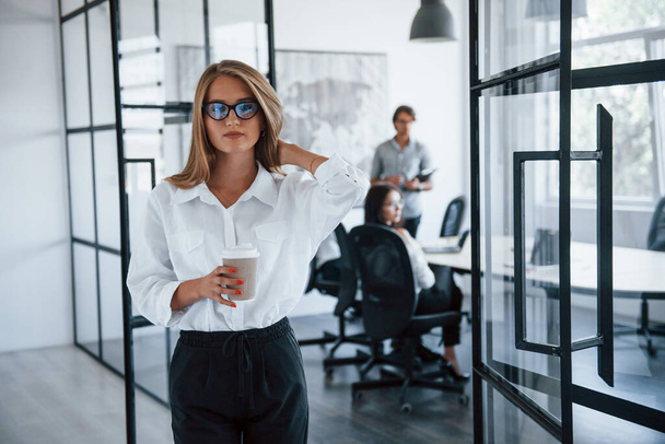 Brunette woman in glasses with cup of drink in hands stands in the office opposite employees. - Φωτογραφία, εικόνα