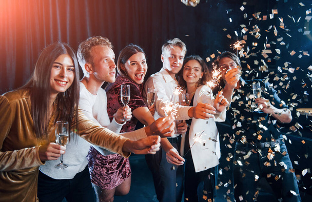 Having fun with sparklers. Confetti is in the air. Group of cheerful friends celebrating new year indoors with drinks in hands. - Photo, image