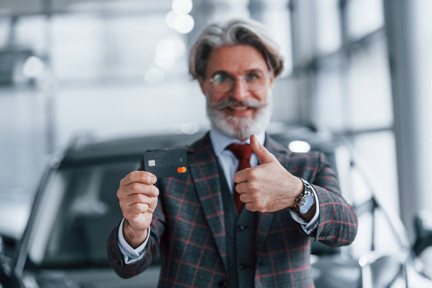 Man with grey hair and mustache standing against modern car indoors with credit card in hand. - Фото, зображення