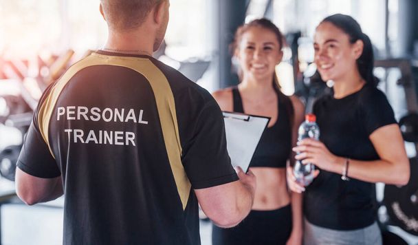 Two young girls in sportive clothes is in the gym together at daytime with their male personal trainer. - Valokuva, kuva