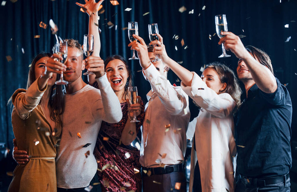 Confetti is in the air. Group of cheerful friends celebrating new year indoors with drinks in hands. - Zdjęcie, obraz
