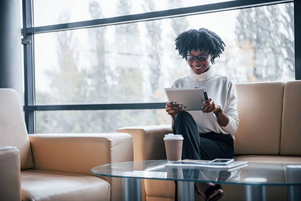 Young african american woman in glasses sits indoors in the office with tablet in hands. - Foto, immagini