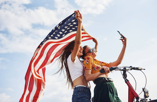 Two patriotic cheerful women with bike and USA flag in hands makes selfie. - Φωτογραφία, εικόνα