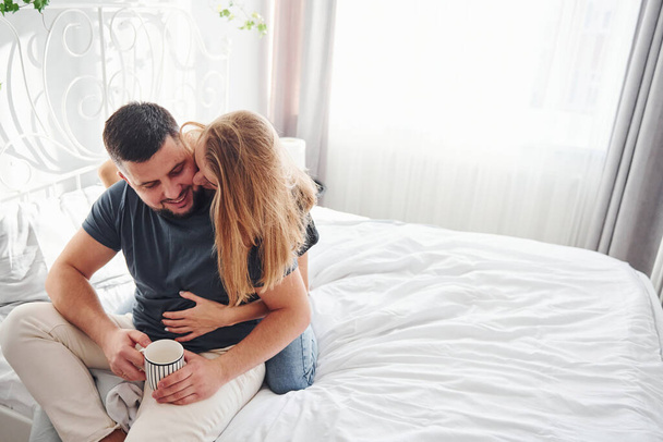 Holding cup of drink. Young married couple have leisure together in bedroom at daytime. - Foto, imagen