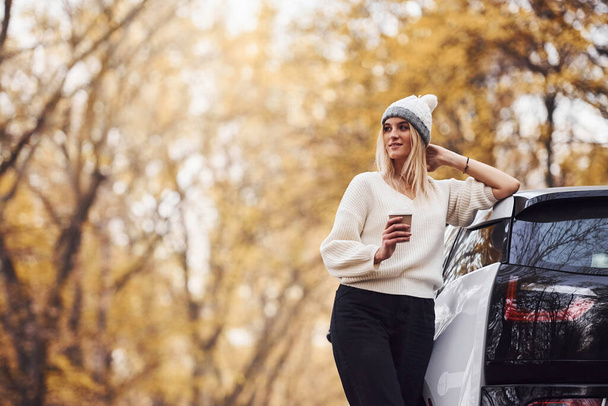 With cup of drink in hands. Girl have autumn trip by car. Modern brand new automobile in the forest. - Photo, image