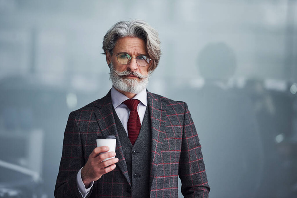 Senior businessman in suit and tie with gray hair and beard standing with cup in hand. - Fotó, kép