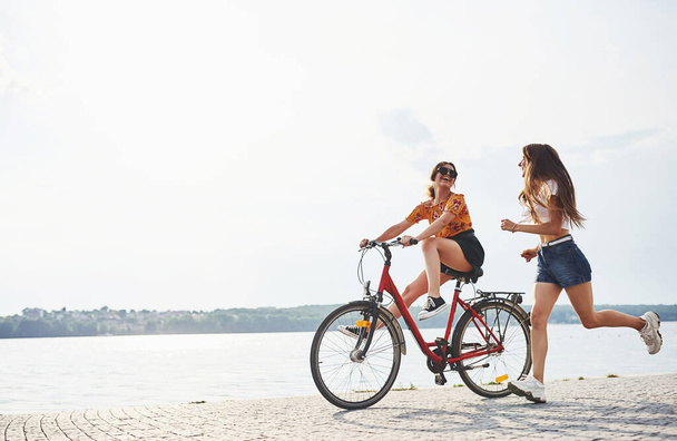 Girl runs near bicycle. Two female friends on the bike have fun at beach near the lake. - Foto, imagen