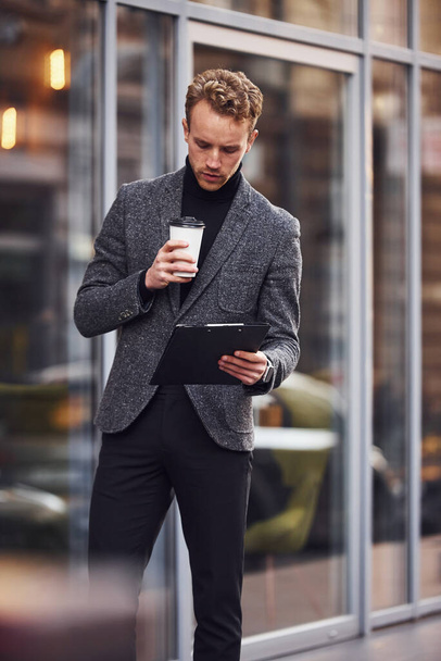 Man in elegant formal wear with cup of drink and notepad in hands is outside against modern building. - Foto, afbeelding