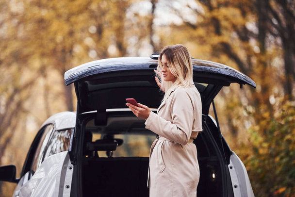 Using smartphone. Girl have autumn trip by car. Modern brand new automobile in the forest. - Fotoğraf, Görsel