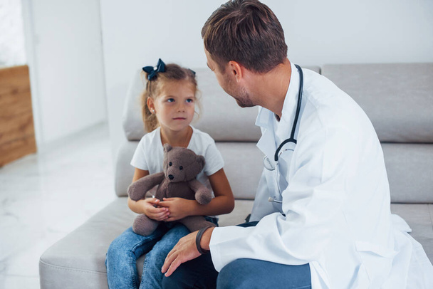 Male doctor in white uniform sits in the clinic with little girl. - 写真・画像