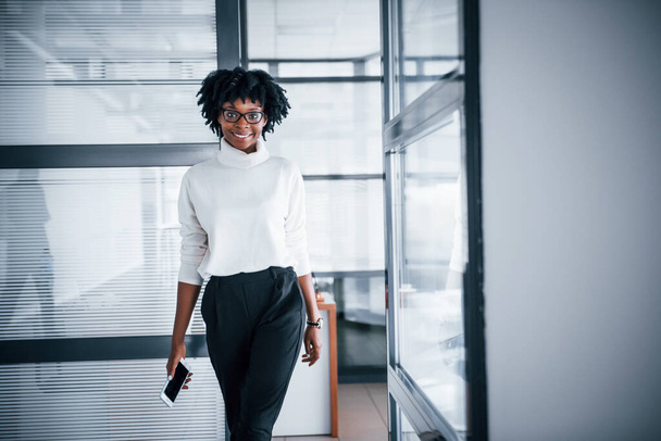Young african american woman in glasses stands indoors in the office with phone. - Photo, Image