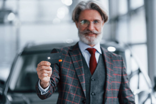 Man with grey hair and mustache standing against modern car indoors with credit card in hand. - Fotó, kép