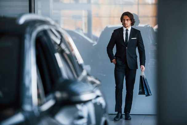Portrait of handsome young businessman in black suit and tie and with shopping bag near the modern car. - Foto, Bild