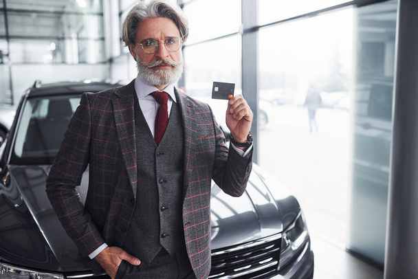 Stylish senior man with gray hair and beard stands against modern black car with credit card in hand. - Fotoğraf, Görsel