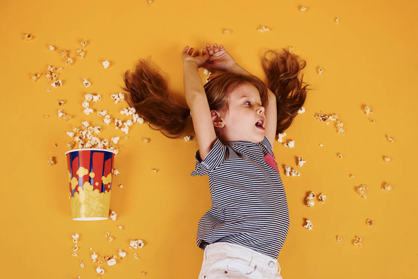 Cute little girl with popcorn lying down on the yellow floor. - Фото, изображение