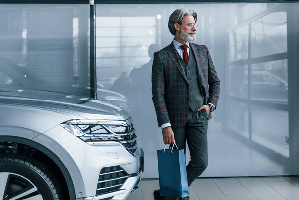 Senior businessman in suit and tie with gray hair and beard standing indoors with blue package near car. - Foto, imagen