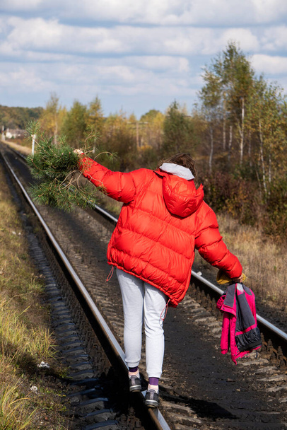 Νεαρή καυκάσια γυναίκα με κυματιστά μαλλιά με κόκκινο μπουφάν που περπατάει στο σιδηρόδρομο σε μικρό χωριό - Φωτογραφία, εικόνα