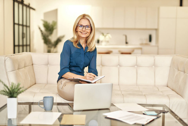 Reife Geschäftsfrau in Freizeitkleidung lächelt in die Kamera und arbeitet von zu Hause aus, während sie in einer modernen Wohnung auf einer Couch vor dem Laptop sitzt - Foto, Bild