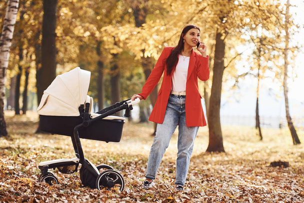 Talking by the phone. Mother in red coat have a walk with her kid in the pram in the park at autumn time. - Foto, immagini