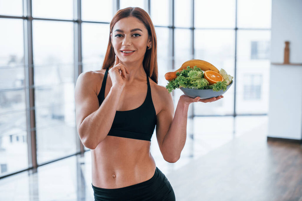 Cheerful sportive woman stands indoors with healthy food in hands. - Foto, Bild
