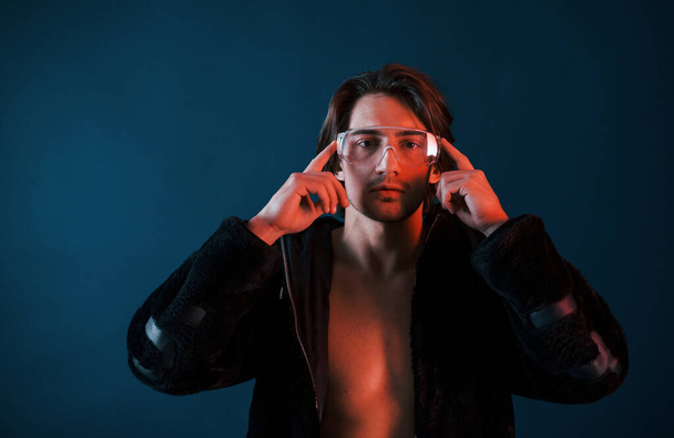 Beautiful long haired hot man in glasses is in the studio with blue neon lighting. - Zdjęcie, obraz