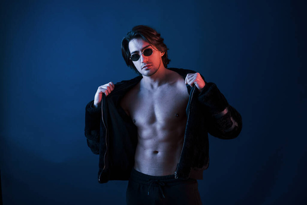 Beautiful man in glasses and black clothes is in the studio with blue neon lighting. - 写真・画像