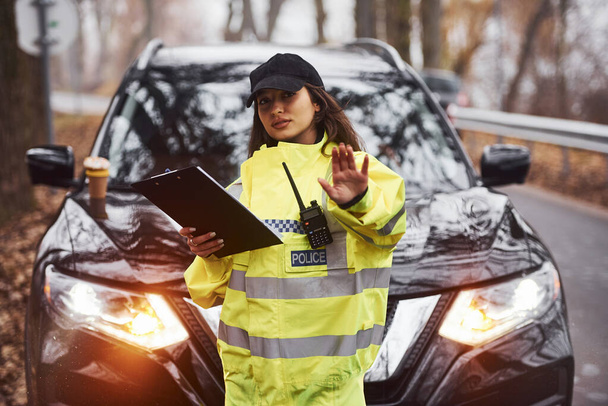 Female police officer in green uniform standing with radio transmitter and notepad against car. - Foto, Imagem