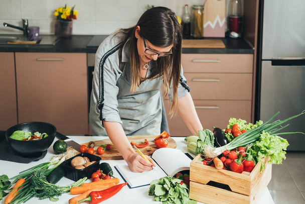 Όμορφη γυναίκα ψάχνει για υγιεινές συνταγές στο βιβλίο μαγειρικής της στο τραπέζι της κουζίνας γεμάτο με φρέσκα λαχανικά για υγιεινό γεύμα - Φωτογραφία, εικόνα