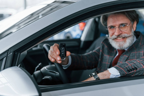Современный стильный пожилой мужчина с седыми волосами и усами, держащий ключи в современном автомобиле. - Фото, изображение
