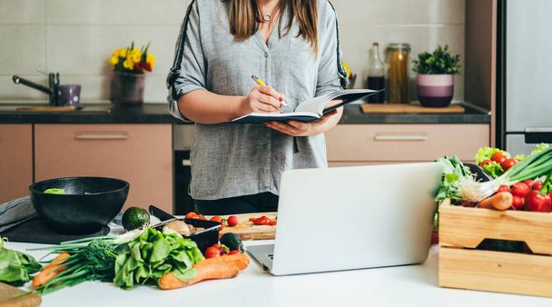 Αγνώριστη γυναίκα γράφει νέα συνταγή σε ένα σημειωματάριο και βλέποντας μαθήματα μαγειρικής σε έναν φορητό υπολογιστή, ενώ στέκεται στο τραπέζι γεμάτο με φρέσκα πολύχρωμα λαχανικά για σπιτικό γεύμα - Φωτογραφία, εικόνα