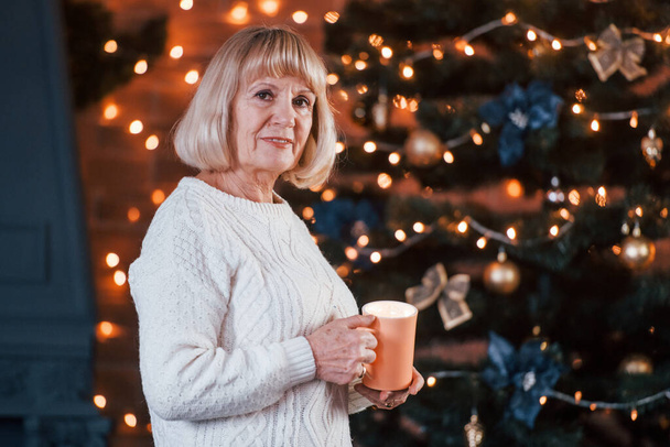 Senior cheerful woman standing with cup of drink in the room with christmas decorations. - Φωτογραφία, εικόνα