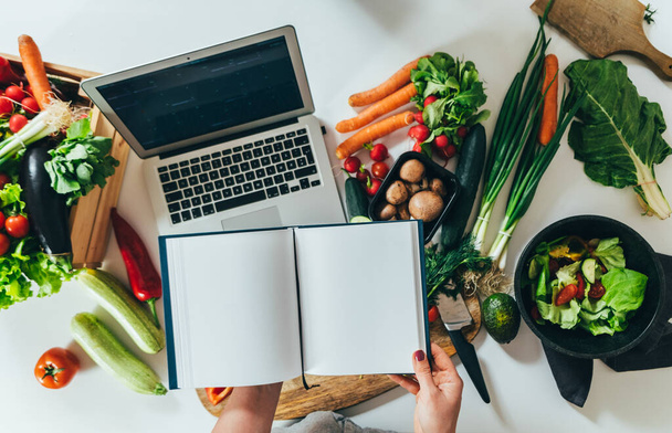 Top view of woman hands holding open blank notebook and watching cooking tutorial on her laptop computer while standing at table full with fresh colorful vegetables and copy space - Фото, зображення
