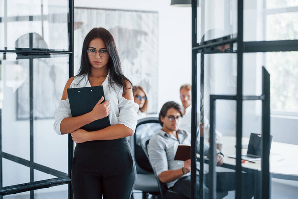 Brunette woman in glasses with notepad in hands stands in the office opposite employees. - Foto, Bild
