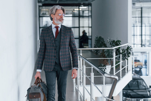Senior businessman in suit and tie with gray hair and beard walking indoors with bag. - Foto, Bild