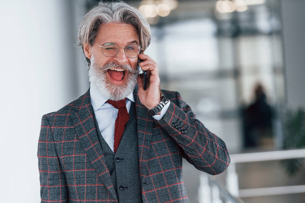 Senior businessman in suit and tie with gray hair and beard standing indoors with phone. - Фото, зображення
