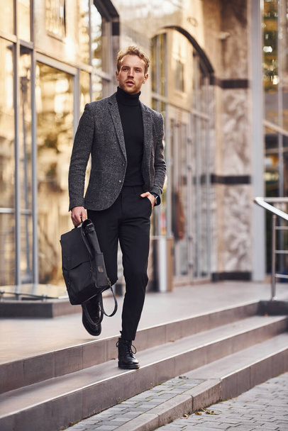 Man in elegant formal wear and with bag is outside against modern building. - Φωτογραφία, εικόνα