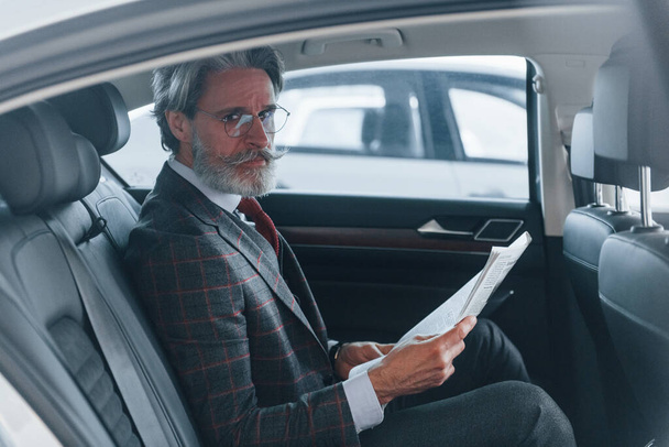 Moderne élégant homme âgé avec les cheveux gris et moustache lecture journal à l'intérieur de la voiture. - Photo, image