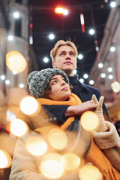 Positive young couple in warm clothes embracing each other on christmas decorated street. - Фото, изображение