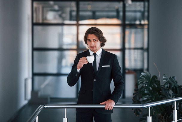 Portrait of handsome young businessman in black suit and tie stands indoors with cup of drink. - Foto, Bild