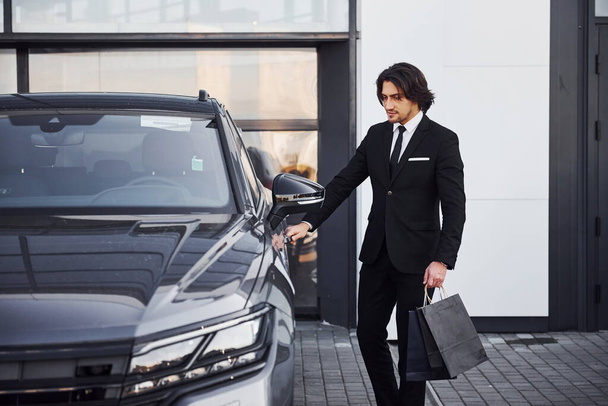 Portrét pohledného mladého podnikatele v černém obleku a kravatě venku v blízkosti moderního auta a s nákupními taškami. - Fotografie, Obrázek