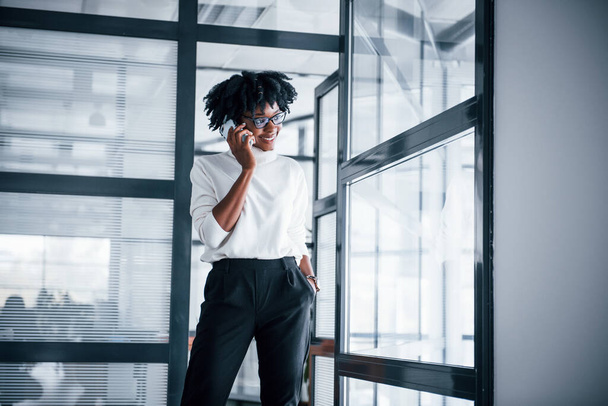 Young african american woman in glasses stands indoors in the office with phone. - Photo, image